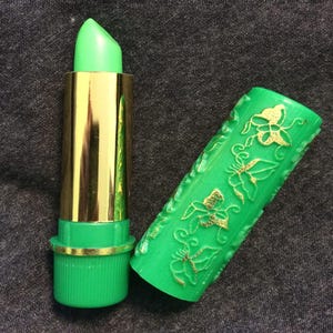 Puede incluir: Un labial verde con un tubo dorado y una base verde a juego. El labial está abierto, revelando el color verde del labial. La tapa verde está decorada con mariposas doradas.