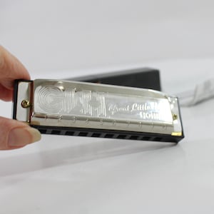 May include: A silver-toned Hohner Great Little Harmonica with a black comb and ten numbered holes. The metal cover plate features the engraved text GLH and Great Little Harmonica. The instrument is held horizontally, showing the front face.