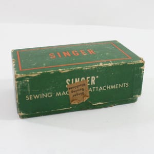 May include: Vintage green Singer sewing machine attachments box. The box has the Singer logo in red and the words "Sewing Machine Attachments" in white. The box is rectangular and shows signs of wear.