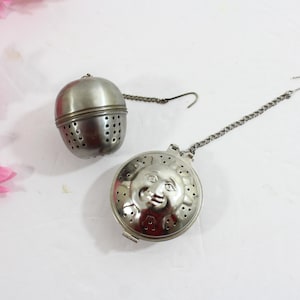 May include: Two silver tea infusers. One is a round, perforated sphere with a chain and hook. The other is a flat, circular infuser with a smiling sun face design, also with a chain and hook. Pink flowers are in the background.