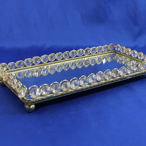 May include: Rectangular vanity tray with a mirrored surface and a gold-colored frame. The frame is adorned with clear, oval-shaped crystals. The tray has small, rounded feet and is set against a blue background.