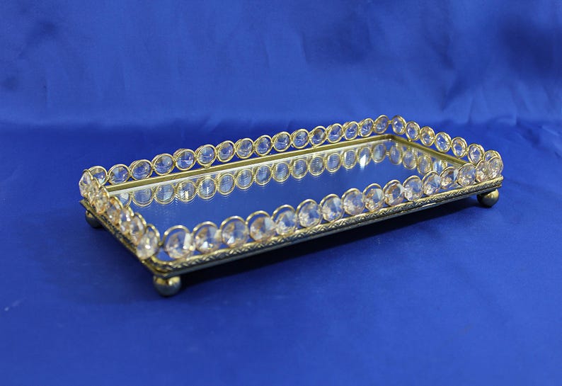 May include: Rectangular vanity tray with a mirrored surface and a gold-colored frame. The frame is adorned with clear, oval-shaped crystals. The tray has small, rounded feet and is set against a blue background.