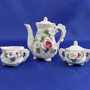 May include: A white porcelain tea set with floral detailing. The set includes a teapot, a creamer, and a sugar bowl. The teapot features a pink rose and other flowers. The set has gold accents and is on a blue background.
