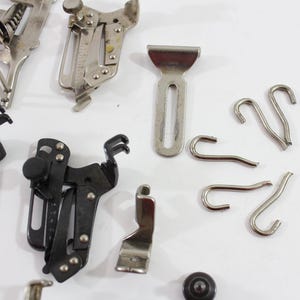 May include: A collection of vintage sewing machine attachments and accessories. The image shows various metal presser feet, hooks, and a small metal plate. The items are in silver and black, and are arranged on a white surface.