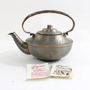 May include: A hammered copper teapot with a silver finish and a matching lid. The teapot has a curved handle and a spout. The teapot is sitting on a white surface with two small cards that say "Gregorian" on them.