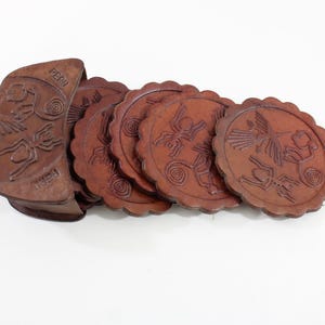 May include: Set of six brown leather coasters with a scalloped edge, featuring embossed designs of stylized figures and animals. The holder is also brown leather and has the word "PERU" embossed on it. Coasters are a home decor item.