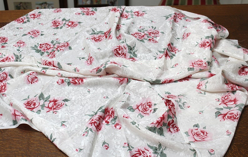 May include: White fabric with a floral pattern of pink and red roses. The fabric is draped and folded.
