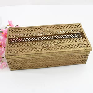 May include: A rectangular, gold-colored metal tissue box with an ornate, openwork design. The box features a hinged lid and decorative floral accents. Pink flowers with green stems are in the background.
