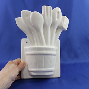 May include: White ceramic kitchen utensil holder featuring a bucket design with wooden texture, holding spoons, a fork, and other cooking tools. The holder is mounted on a rectangular plaque with a hole for hanging. The background is a solid blue.