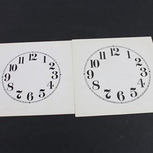 May include: Two white clock face templates with black numerals and markings. The numbers 1 through 12 are printed around the circular face. The templates are on a black surface.