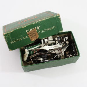 May include: Vintage green Singer sewing machine attachments in a green box. The box is labeled "SINGER" and "SEWING MACHINE ATTACHMENTS". The box is open, revealing various metal attachments.