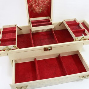 May include: Cream-colored jewelry box with red velvet lining and multiple compartments. The box has a hinged lid with a gold floral design and several pull-out trays. The box is open, revealing its interior and storage spaces.
