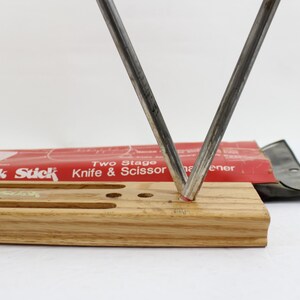 May include: A knife and scissor sharpener with a wooden base and two metal sharpening rods in a V-shape. The red packaging reads "Crock Stick Knife & Scissor Sharpener".