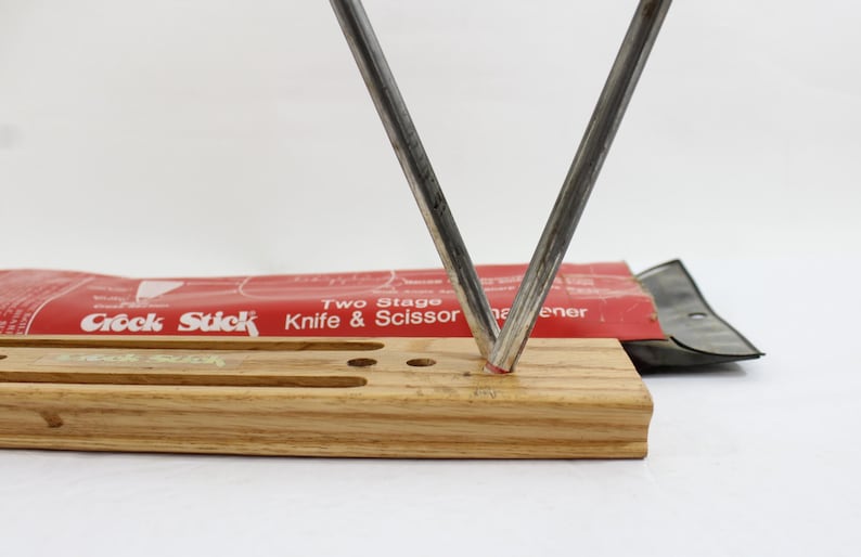 May include: A knife and scissor sharpener with a wooden base and two metal sharpening rods in a V-shape. The red packaging reads "Crock Stick Knife & Scissor Sharpener".