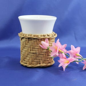 May include: A white ceramic cup with a woven brown wicker cup holder. Pink flowers are in the foreground.