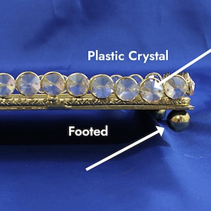 May include: A gold-toned, footed tray with a decorative border of clear plastic crystals. The tray is rectangular and sits on small, round feet. The crystals are set in a repeating pattern along the edge.