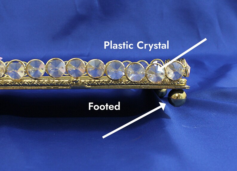 May include: A gold-toned, footed tray with a decorative border of clear plastic crystals. The tray is rectangular and sits on small, round feet. The crystals are set in a repeating pattern along the edge.