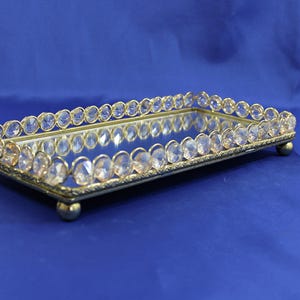May include: A rectangular decorative tray with a mirrored surface and a gold-colored frame. The frame is adorned with clear, oval-shaped crystals. The tray rests on four small, gold-colored ball feet, creating an elegant display.