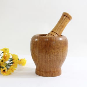 Puede incluir: Un mortero y un pilón de madera. El mortero es marrón y tiene una forma redondeada. El pilón también es marrón y tiene un mango liso y redondeado.