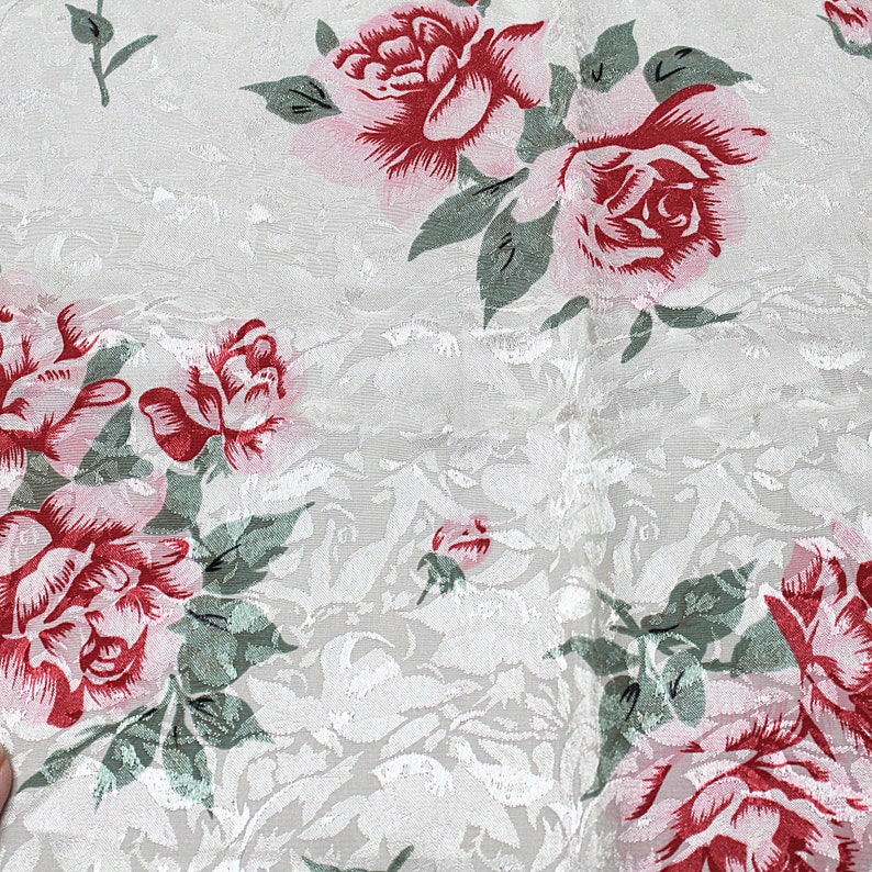 May include: White fabric with a repeating pattern of pink and red roses. The roses have a textured appearance and are surrounded by green leaves.