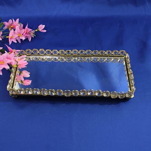 May include: A rectangular mirrored tray with a gold-colored frame and clear crystal-like accents. The tray is on a blue surface, and pink flowers are in the upper left corner.