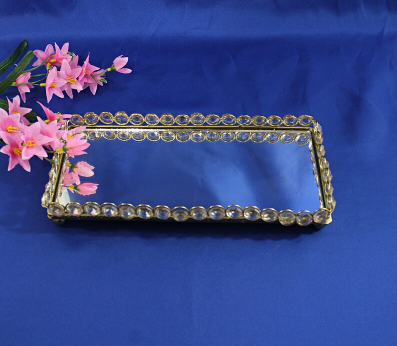 May include: A rectangular mirrored tray with a gold-colored frame and clear crystal-like accents. The tray is on a blue surface, and pink flowers are in the upper left corner.