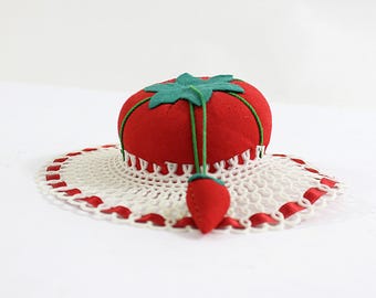 May include: A red felt tomato pincushion with a green felt leaf on top, sitting on a white crocheted doily with red ribbon trim.