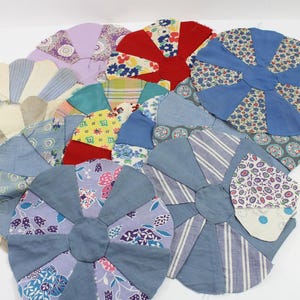 May include: A collection of handmade, circular fabric items in various colors and patterns. The pieces feature a pinwheel design with shades of blue, red, purple, and cream. The fabrics include floral prints, stripes, and solid colors, creating a patchwork effect.
