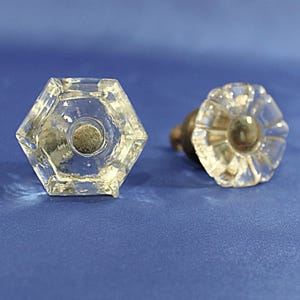 May include: Two clear glass cabinet knobs. One is hexagonal, the other is a flower shape. Both have a dark center and brass hardware. The knobs are on a blue background.