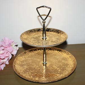 May include: Two-tiered gold serving stand with a hammered finish. The stand has a silver handle and is perfect for displaying desserts or snacks.