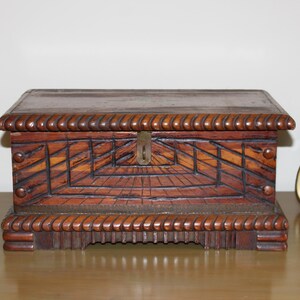 May include: A small, rectangular wooden box with a hinged lid. The box is dark brown with intricate geometric carvings on the front and sides. The edges are adorned with a rope-like design. A small metal clasp secures the lid.