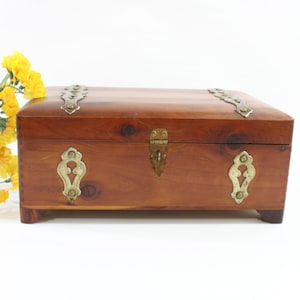 May include: A rectangular wooden chest with brass hardware, including corner accents, a latch, and decorative escutcheons. The chest is a warm brown color and sits on small feet. Yellow flowers are in the background.