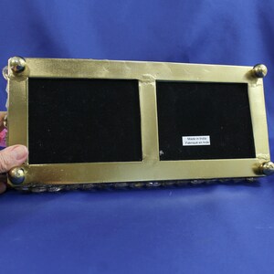 May include: A rectangular, gold-colored picture frame with two openings, each with a black velvet backing. The frame has rounded corners and small, gold ball feet. The bottom edge is lined with small, clear, decorative stones. The text "Made in India Fabriqu&eacute; en Inde" is visible.