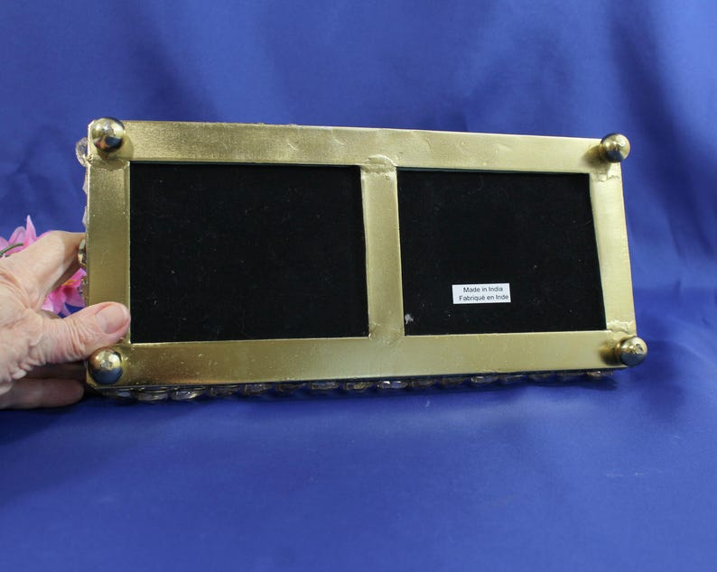 May include: A rectangular, gold-colored picture frame with two openings, each with a black velvet backing. The frame has rounded corners and small, gold ball feet. The bottom edge is lined with small, clear, decorative stones. The text "Made in India Fabriqu&eacute; en Inde" is visible.