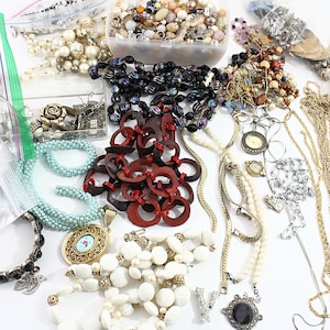 May include: A collection of various jewelry pieces, including necklaces, bracelets, and earrings, in a variety of colors and materials. There are beaded necklaces, gold chains, and a red wooden necklace. Some items are in plastic bags and containers.