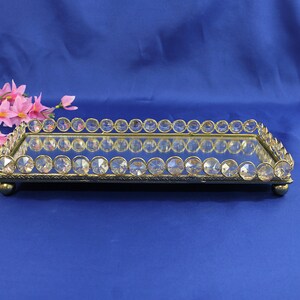 May include: A rectangular, mirrored tray with a gold-colored metal frame. The frame is adorned with clear, round crystals along the edges. The tray sits on four small, rounded feet. Pink flowers are visible in the background.