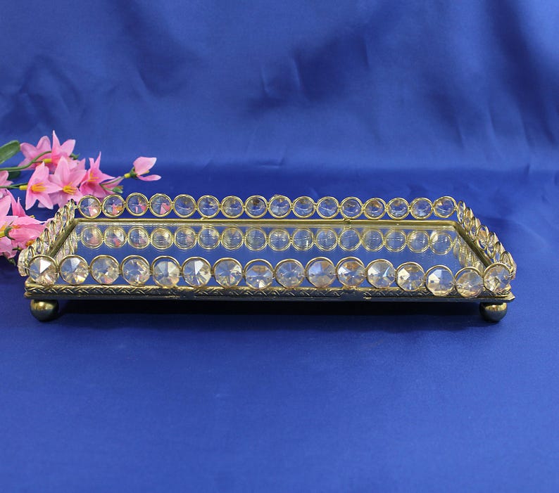 May include: A rectangular, mirrored tray with a gold-colored metal frame. The frame is adorned with clear, round crystals along the edges. The tray sits on four small, rounded feet. Pink flowers are visible in the background.