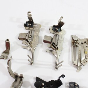 May include: A collection of vintage sewing machine presser feet in various shapes and sizes. The feet are made of silver-colored metal and are arranged on a white surface. Some feet have intricate designs and levers.