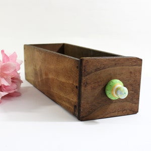May include: A rectangular wooden box with a decorative knob. The box is stained a warm brown color. The knob is green, yellow, and white. Pink flowers are in the background.