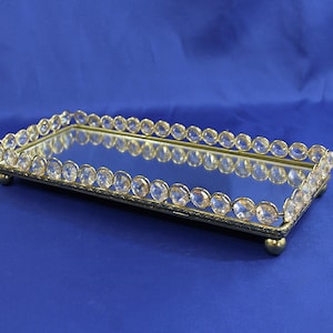 May include: Rectangular mirrored tray with a gold-colored metal frame. The frame is adorned with clear, round crystals. The tray sits on small, rounded feet. The background is a solid blue.