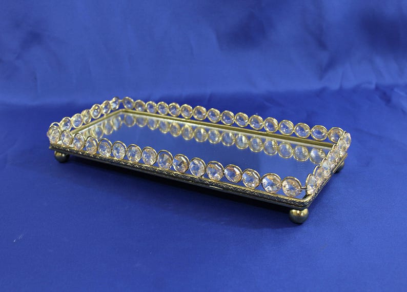 May include: Rectangular mirrored tray with a gold-colored metal frame. The frame is adorned with clear, round crystals. The tray sits on small, rounded feet. The background is a solid blue.