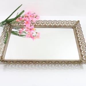 May include: A rectangular vanity tray with a mirrored surface and an ornate, filigree-style metal frame. The frame has a gold finish. Pink flowers with green stems are placed on the tray.