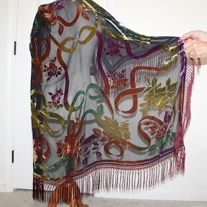 May include: A sheer black shawl with colorful velvet floral and ribbon designs. The shawl features a fringe trim in a deep burgundy color. The ribbons are in shades of orange, green, and gold. The flowers are in shades of purple, red, and yellow.
