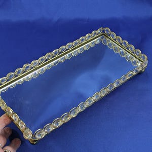 May include: Rectangular mirrored tray with a gold-colored frame. The frame is adorned with clear, oval-shaped crystals. The tray sits on small feet and is displayed against a blue background. A hand is visible on the left side.