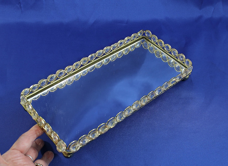May include: Rectangular mirrored tray with a gold-colored frame. The frame is adorned with clear, oval-shaped crystals. The tray sits on small feet and is displayed against a blue background. A hand is visible on the left side.
