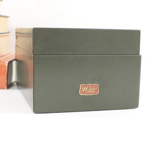 May include: A rectangular olive green metal storage box with a hinged lid. The front of the box features a small, decorative red and gold label with the text "Weir" in a stylized script font. The box has a smooth, matte finish.