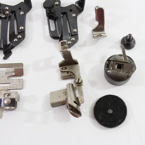 May include: A collection of vintage sewing machine parts, including presser feet, a bobbin case, and other metal components. The parts are in various shades of silver and black, with some pieces bearing the "SINGER USA" mark.