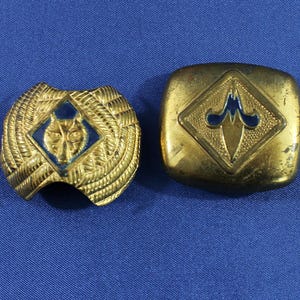 May include: Two vintage gold-toned metal pins with blue enamel accents. One pin is a shield shape with a wolf's head, the other is a square shape with a fleur-de-lis design. Both are on a blue background.