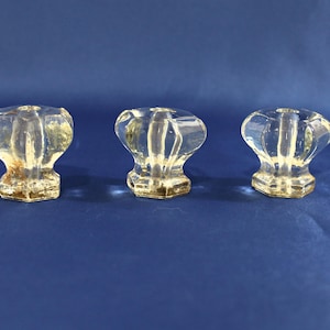 May include: Three clear glass cabinet knobs with a ribbed, rounded top and a square base. The knobs are arranged in a row against a blue background. The glass has a slight yellow tint.