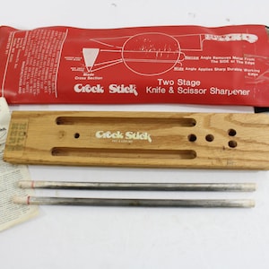 May include: A Crock Stick two-stage knife and scissor sharpener kit. The kit includes a wooden base with the text "Crock Stick", two sharpening rods, a red pouch with instructions, and a small instruction sheet. The pouch reads "Two Stage Knife & Scissor Sharpener".